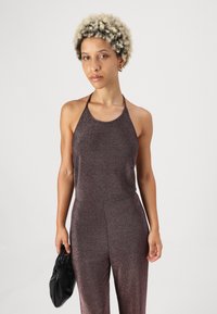 Mono metálico brillante y oscuro con cuello halter y forma ajustada. Combinado con un bolso negro anudado, el tejido tiene una textura lisa y reflectante.
