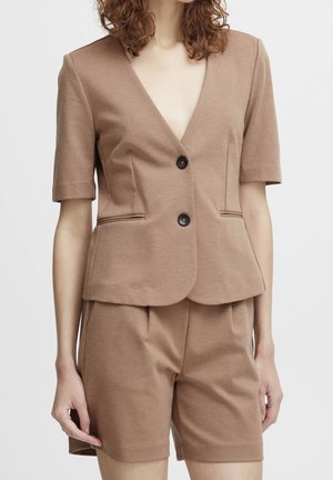 Kvinde iført en tan kortærmet blazer med to knapper og matchende skræddersyede shorts mod en ensfarvet baggrund.