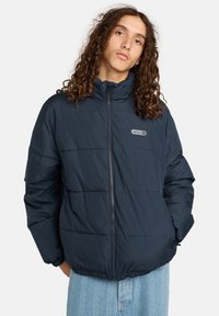 Giovane con lunghi capelli ricci che indossa un giubbotto imbottito blu navy con zip frontale e jeans a motivi blu chiaro.