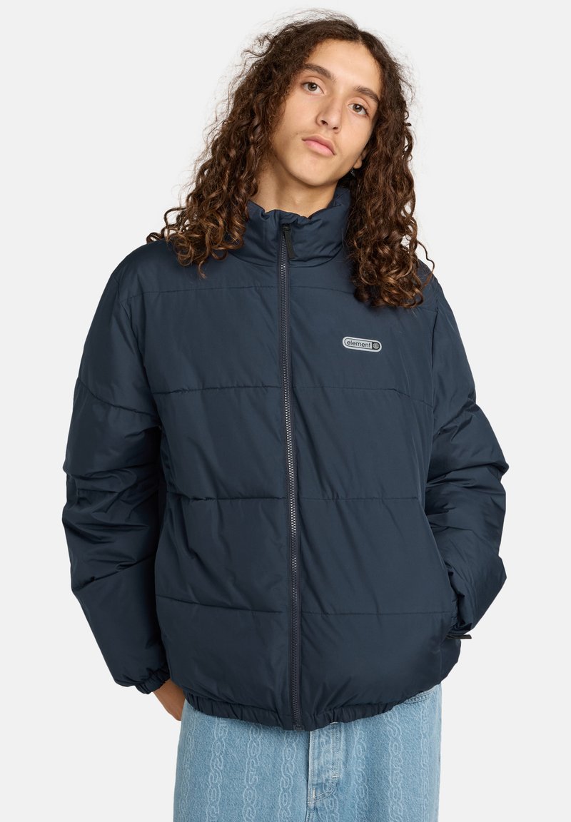 Giovane con lunghi capelli ricci che indossa un giubbotto imbottito blu navy con zip frontale e jeans a motivi blu chiaro.