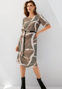 Kleid mit Gürtel, das abstrakte braune und schwarze Muster aufweist, kurze Ärmel hat und knielang geschnitten ist. Tragen mit schwarzen Knöchelstiefeln.