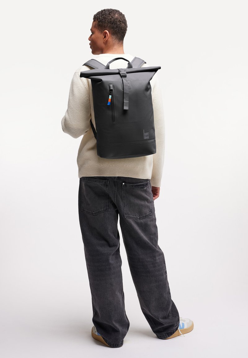 Joven vistiendo un suéter color crema y jeans negros, llevando una mochila enrollable negra con cremallera frontal y tirador de cremallera colorido, visto desde atrás.