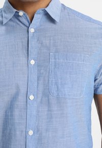 Jasnoniebieska koszula na krótki rękaw z białymi guzikami i lewą kieszenią na piersi, noszona przez osobę przedstawioną od szyi i ramienia.