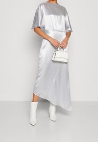 Robe midi en satin argenté avec des manches courtes flottantes, taille cintrée et ourlet asymétrique, assortie à des bottines blanches et un sac à main.