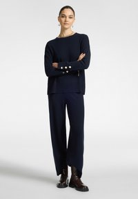 Merihenkinen neulepusero, jossa on pitkät hihat ja kultaiset napit, yhdistettynä leveälahkeisiin merihenkisiin housuihin ja ruskeisiin nilkkurit. Pintamateriaali on sileä ja muotoilu on rento.