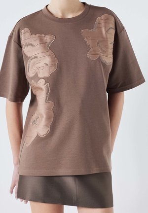 Brown loose short-sleeve shirt with shiny floral appliques worn over a dark brown skirt, shown on a person from neck to mid-thigh.