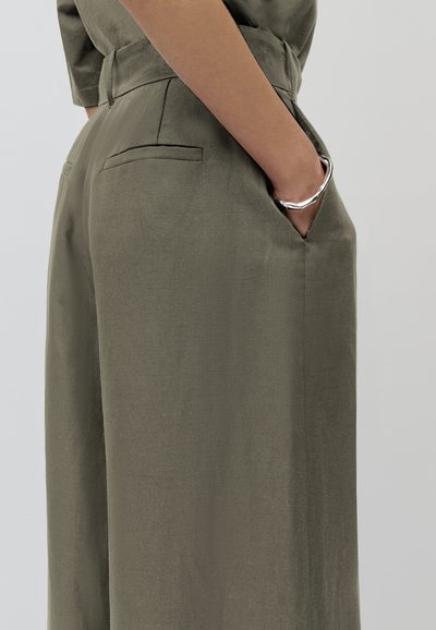Olivengrüne weite Beinhosen aus strukturierter Stoff, mit hoher Taille, Seitentaschen und schmalem Schnitt an den Knöcheln. Sichtbares Detail mit silbernem Ring.