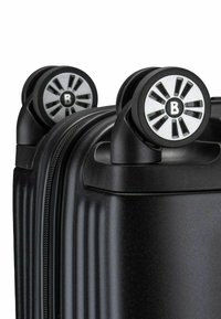 Valigia rigida nera con texture a coste verticali, dotata di due ruote nere e argento con motivo circolare e chiusura con cerniera.