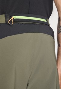 Olivengrønne treningsshorts med svart livintervall, med en glidelåslomme med limegrønn glidelås og oransje drahåndtak. Jevn tekstur.