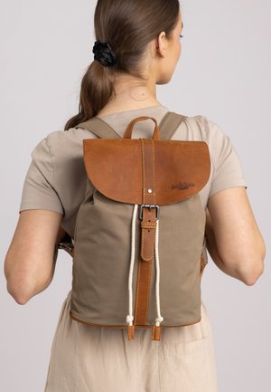 Canvas backpack in khaki with a brown leather flap, leather strap, and rope drawstring. Features a top handle and prominent stitching detail.