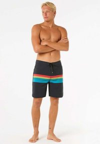 Shorts de baño negros con rayas horizontales de arcoíris, cordón ajustable y una textura de tela suave. Llevados por una persona de pie descalza.
