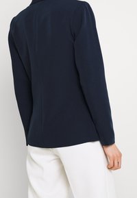 Person iført en tætsiddende marineblå blazer og løse hvide bukser, set bagfra mod en ensartet lys baggrund.