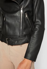 Giacca da motociclista in pelle nera con zip argentate, vita con fibbia e maniche testurizzate; vista ravvicinata della parte anteriore e della mano.