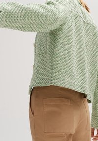 Grön texturerad jacka med croppad design, med ett vävt mönster och två knappar, matchad med beige byxor med bakfickor.