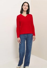 Maglione rosso lavorato a maglia con scollo a V, orlo a coste e maniche lunghe, abbinato a jeans svasati blu e sneakers bianche. Design semplice, tessuto testurizzato.