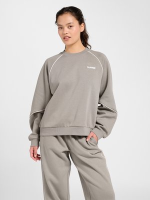 Ung kvinde iført lys grå Hummel sweatshirt og matchende joggingbukser, stående med den ene hånd i lommen mod en ensfarvet baggrund.