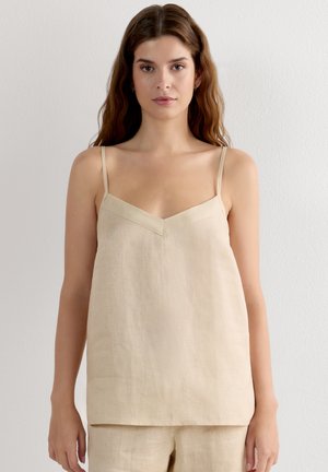 Vrouw met lang bruin haar die een beige mouwloze linnen top met V-hals en bijpassende broek draagt, staand tegen een witte muur.