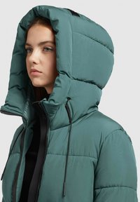 Jachetă verde teal căptușită, cu o glugă mare, având fermoar și șnururi ajustabile. Textură netedă cu detalii de quilt.