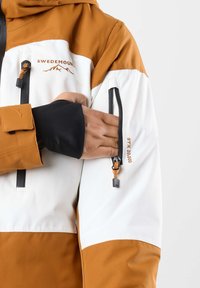 Giacca impermeabile con la parte superiore di colore marrone e la parte inferiore di colore bianco. Include tasche con zip e polsini regolabili con materiali elasticizzati.