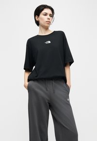 Νεαρό άτομο που φοράει μαύρο μπλουζάκι The North Face και γκρι παντελόνι με κορδόνι, με τα χέρια στις τσέπες, κοιτάζοντας ελαφρώς προς τα αριστερά πάνω από απλό φόντο.