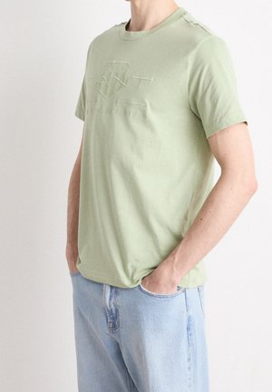 Camiseta de algodón verde claro con cuello redondo, mangas cortas y detalle de logotipo en relieve en la parte delantera. Combinada con jeans de mezclilla azul claro.