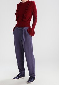 Karmesinroter Pullover mit gerafftem Vorderteil, kombiniert mit lockeren, dunkelblauen Hosen. Das Modell trägt glänzende navyfarbene Sneakers. Schlichtes, modernes Design.