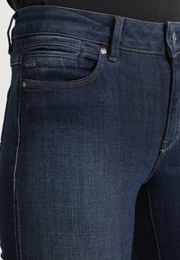 Jeans de mezclilla azul oscuro con diseño de cinco bolsillos, costuras clásicas y talle medio. La tela presenta una textura suave con un ligero desvanecimiento.