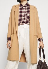 Camel-wollemantel mit offenem Vorderteil, seitlichen Taschen und aufgerollten Ärmeln über einem karierten bordeauxfarbenen Hemd mit Knöpfen und hellen Leinenhosen.