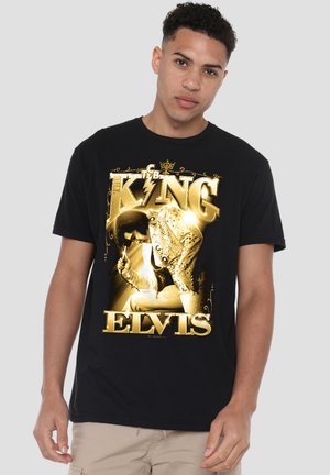 Young man wearing a black T-shirt with a gold graphic of Elvis Presley and text "King Elvis" on front, standing against a plain light background.