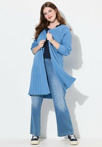 Hellblauer, gerippter Cardigan mit langen Ärmeln, offenem Vorderteil und plissiertem Design. Kombiniert mit einem dunklen Oberteil und ausgestellten Jeans, weißen Sneakers.
