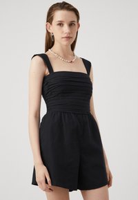 Mono de lino negro con escote cuadrado estructurado, cuerpo fruncido y tirantes anchos. Presenta pantalones cortos sueltos y una textura suave.