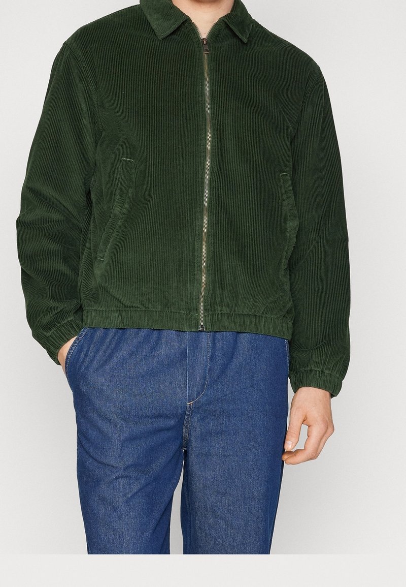 Donkergroene corduroyjas met een rits, kraag, zijzakken en elastische manchetten. Gecombineerd met blauwe denim broek.