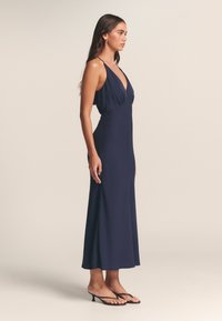 Moteris dėvi tamsiai mėlyną midi suknelę be rankovių ir su plonomis petnešėlėmis, stovinti profiliniu kampu ant neutralios spalvos fono, avinti juodus aukštakulnius sandalutus.