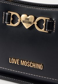 Svart läderhandväska med guldfärgad hårdvara, hjärtformade detaljer och varumärkestexten "LOVE MOSCHINO" präglad i guld längst ner.