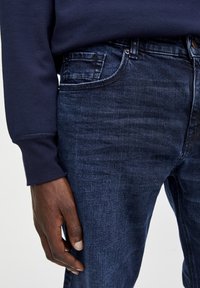 Mörkblå denimjeans med en texturerad yta, som har en standard femfickorsdesign och en synlig midjebandknapp.