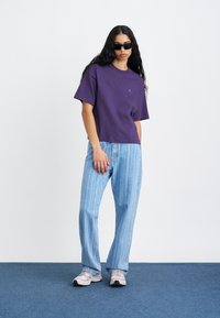 Violeta kokvilnas t-krekls ar īsām piedurknēm un mazu logotipu, apvienots ar gaiši ziliem vertikāli svītrainiem džinsiem un pelēkiem sporta apaviem.