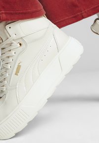 Białe sneakersy typu high-top z złotymi akcentami, z teksturowaną skórzaną cholewką, grubą białą podeszwą i widocznym logo na boku.