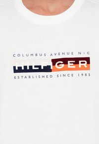 Hvit skjorte med "Columbus Avenue NYC," en fargeblokkerte "HILFIGER" logo, og teksten "Etablert siden 1985" på brystet.