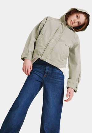 Giovane con giacca con cappuccio verde chiaro e jeans blu a gamba larga, inclinato leggermente da un lato contro uno sfondo semplice.