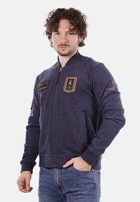 Giacca bomber blu navy con zip frontale, colletto a coste e toppe in stile militare su petto e manica. Tessuto morbido, vestibilità casual.