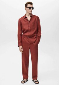 Red linen shirt with a collar and pocket, paired with matching loose-fit trousers and black flip-flops. Model wearing dark sunglasses.