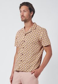 Camisa de manga corta de tela rosa claro con un patrón geométrico de zig zag en tonos de amarillo, rojo y marrón. Con cierre de botones al frente.