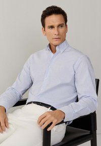 Muškarac sjedi na crnoj stolici, nosi bijele hlače i svijetloplavu košulju s uzorkom na kockice, gleda blago prema desnoj strani.