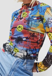 Long-sleeve mesh top features a colorful graffiti print with smiley faces, flowers, and logos. Lightweight fabric, fitted design, scalloped hem.
