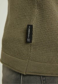 Oliwkowy sweter z teksturowanej tkaniny, z prążkowanym końcem i etykietą po boku. Materiał wydaje się miękki z wzorem splotu.