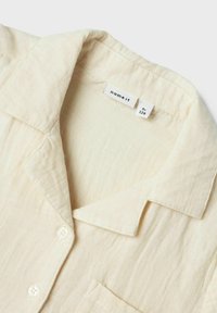 Camisa de color crema, de manga corta, con una tela suave y texturizada. Presenta un collar de solapa, dos botones y un bolsillo en el pecho.