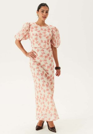 Kvinna står och bär en vit maxiklänning med rosa blommönster och puffade korta ärmar, kombinerad med mörka klackar och minimal smyckning.