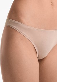 Beige tanga gemaakt van zachte, rekbare stof. Heeft een dunne tailleband en een ontwerp met minimale bedekking, met een gladde textuur en zonder zichtbare patronen.