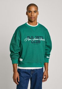 Sudadera verde con cuello redondo, que presenta el texto "PJL 1973 Pepe Jeans London CLUB" en azul marino y blanco. Tejido suave y corte relajado.
