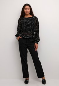 Blusa nera a pois con maniche lunghe, vita smock, abbinata a pantaloni neri e scarpe con punta. Tessuto liscio e trasparente.